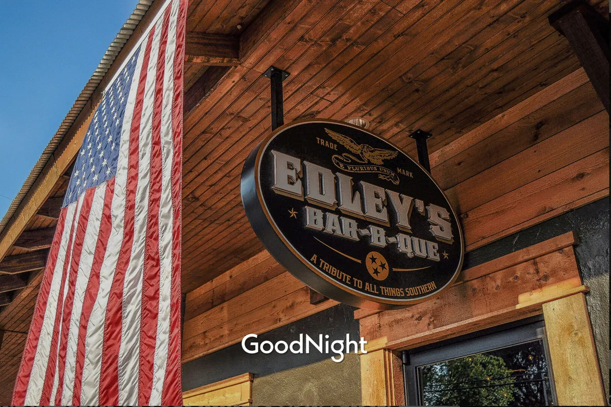This image features the iconic Edley's Bar-B-Que with its recognizable signage and American flag, set against a welcoming exterior. The scene suggests an inviting atmosphere for both locals and visitors to gather and enjoy classic Southern cuisine, with hints of community events and live music nights.
