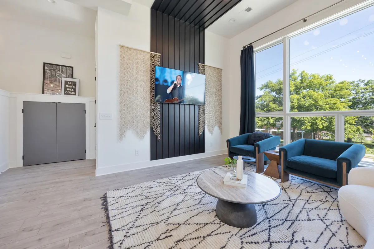 A spacious second living area with a modern black feature wall housing a mounted television, blue accent chairs, a stylish coffee table, and a large window that allows natural light to fill the space. The room is arranged for both comfort and entertainment, ideal for movie nights or casual gatherings.