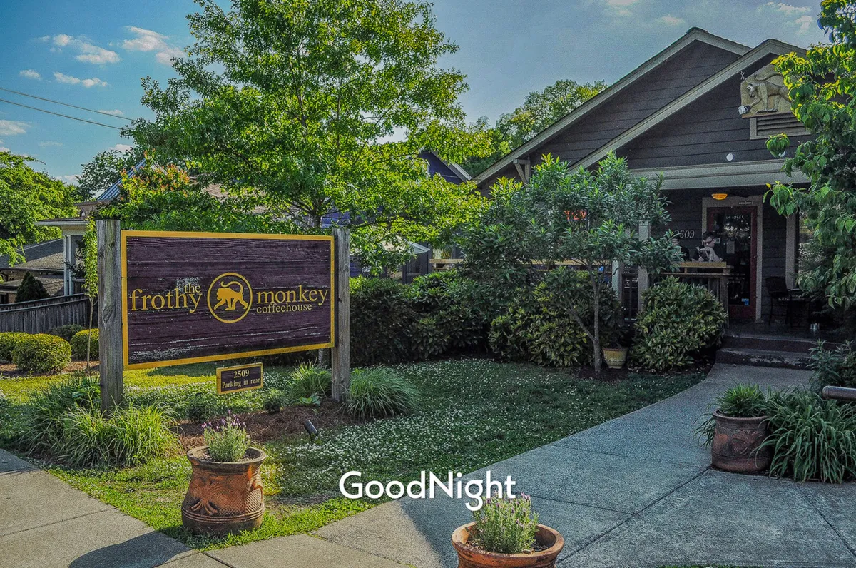 A friendly, welcoming exterior shot of The Frothy Monkey coffeehouse, featuring its inviting signage, landscaped garden, and bustling entrance. The scene conveys the social energy of a beloved local spot, where guests can enjoy coffee or mingle outdoors.