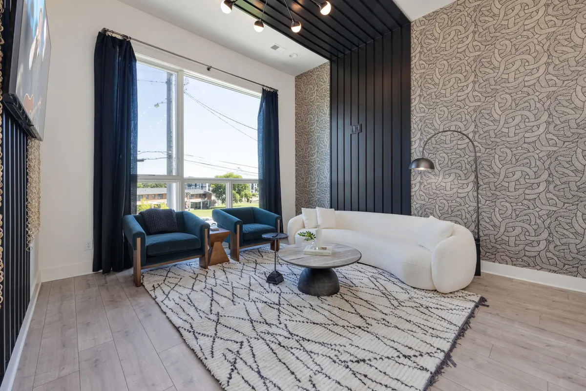 A modern living room with a plush white sofa and two teal armchairs set around a decorative rug. A large window brings in natural light, while a floor lamp adds a cozy ambiance. This space is ideal for unwinding, entertaining, or enjoying relaxed conversations.
