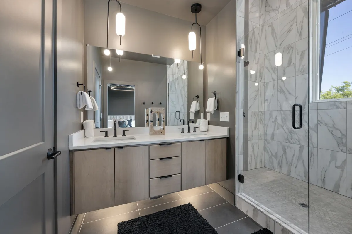 A serene and contemporary bathroom with a dual sink vanity, large mirror, glass shower enclosure, and elegant hanging light fixtures. The space feels bright and refreshing, ideal for morning routines or unwinding after a busy day.