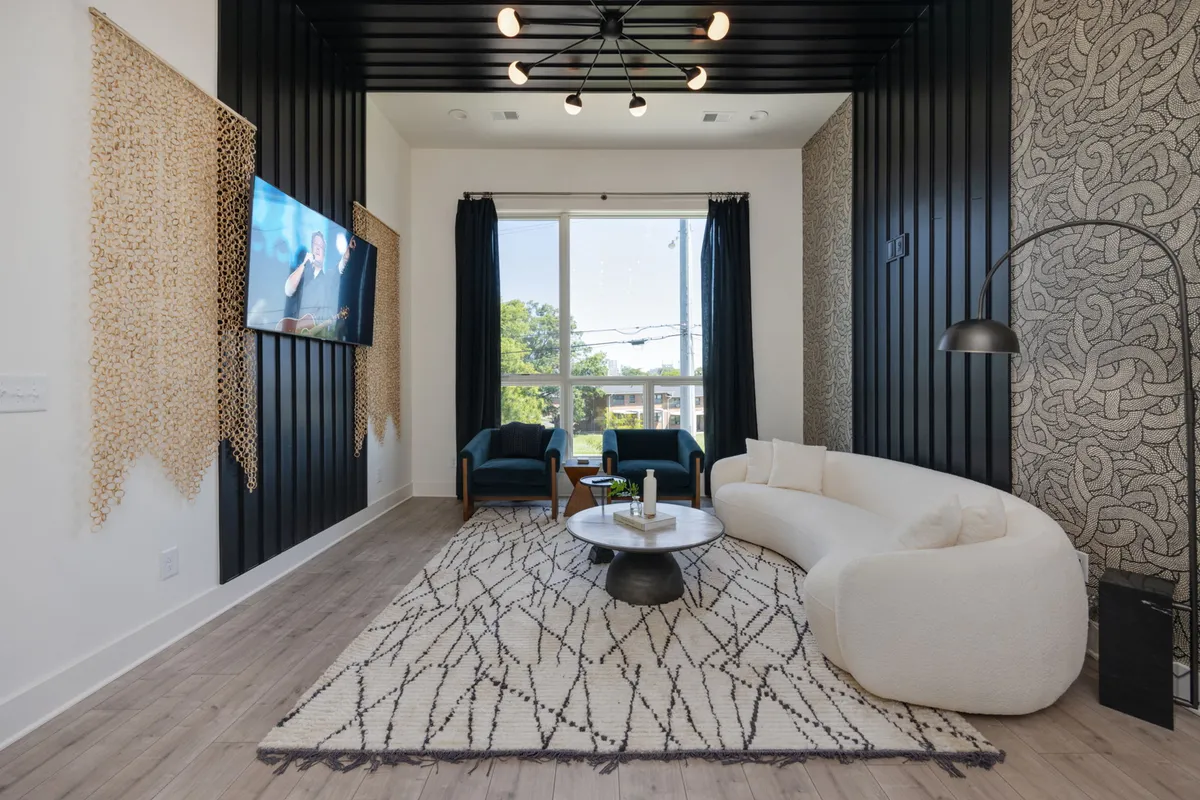 The image showcases a stylish, contemporary living room with a striking curved white sofa set against a dramatic black accent wall. Sunlight pours in from a large window, illuminating a textured wall hanging and a chic coffee table, creating a space designed for relaxation, conversation, and cozy movie nights.