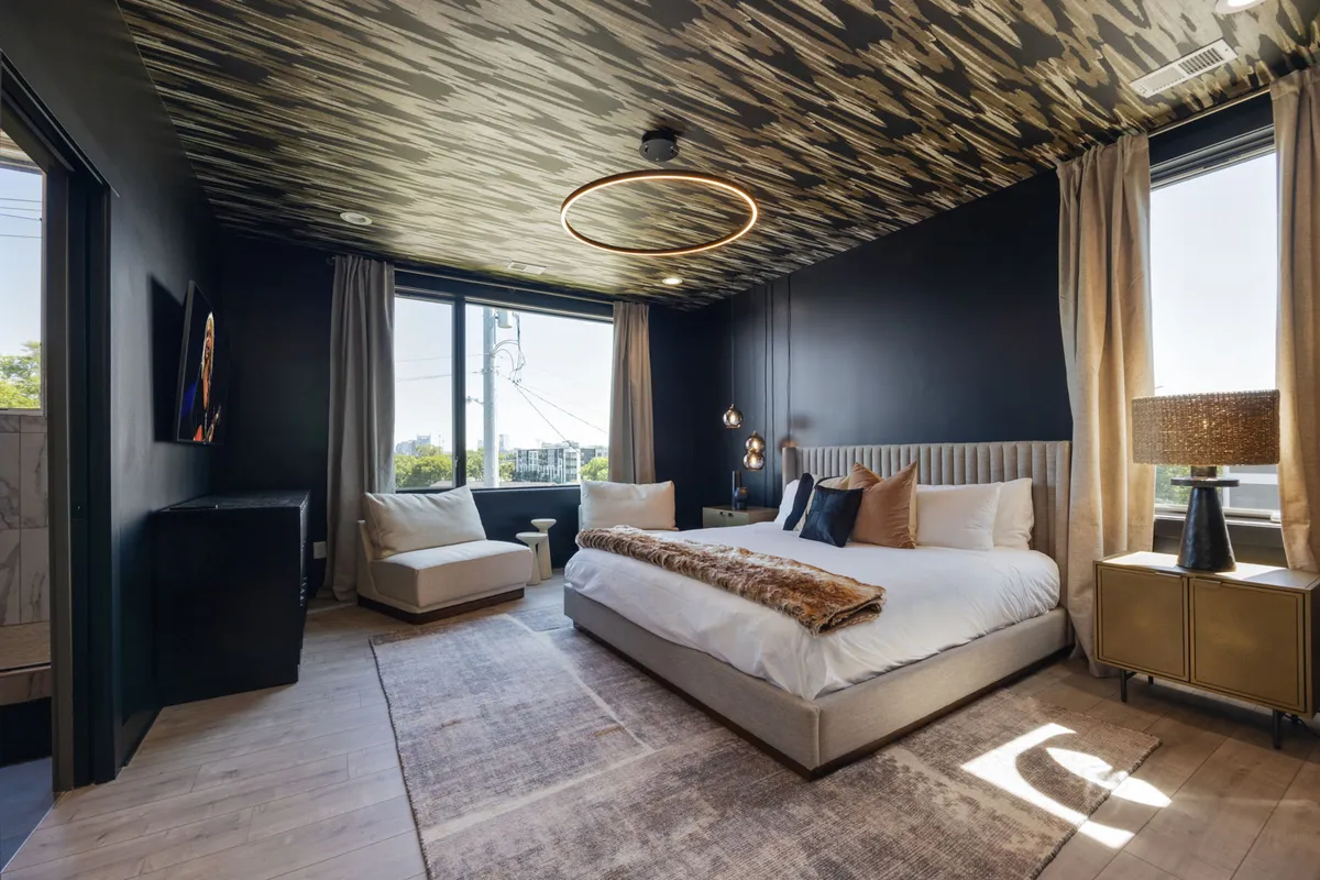 A spacious, sunlit bedroom and living area featuring a plush bed, cozy sofa, elegant lighting fixtures, and large windows that fill the room with natural light. The inviting setup suggests a comfortable, relaxing environment with a touch of sophistication.
