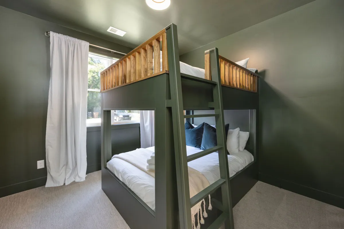 A playful yet tranquil bedroom featuring queen bunk beds with textured pillows and a window with flowing curtains. The arrangement suggests a welcoming space for sleepovers, family stays, or friends traveling together, with an atmosphere that balances relaxation and fun.