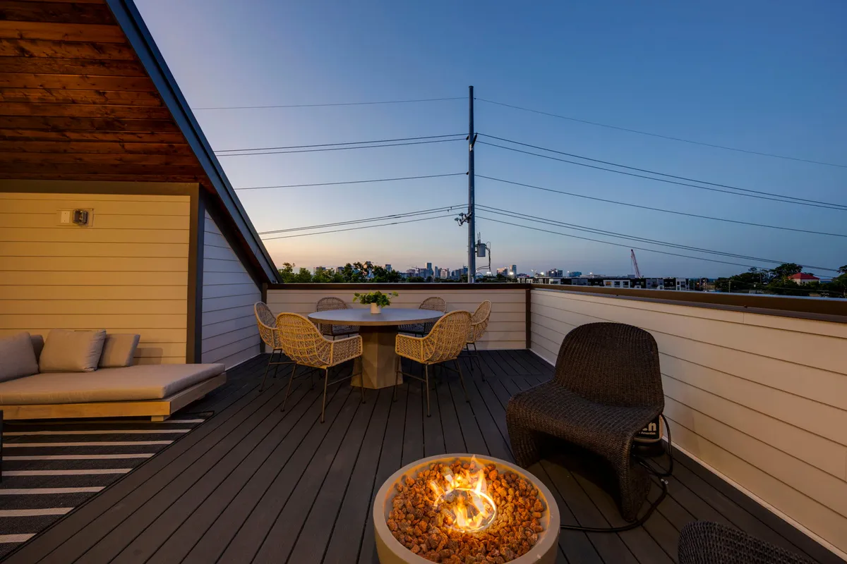 An elegant rooftop scene with a fire pit, dining table, lounge seating, and the city skyline in the background at sunset. The warm glow of the fire and ambient lighting create an inviting atmosphere for social gatherings and intimate evenings.