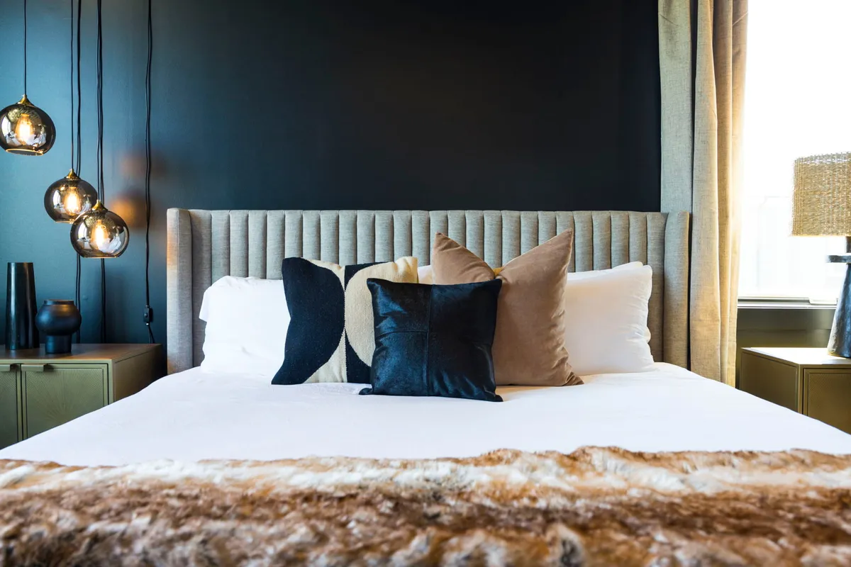 A tranquil bedroom scene with a beautifully made bed adorned with decorative pillows, ambient pendant lighting, a textured headboard, and stylish nightstands. The space feels like a peaceful sanctuary, ideal for restful nights.