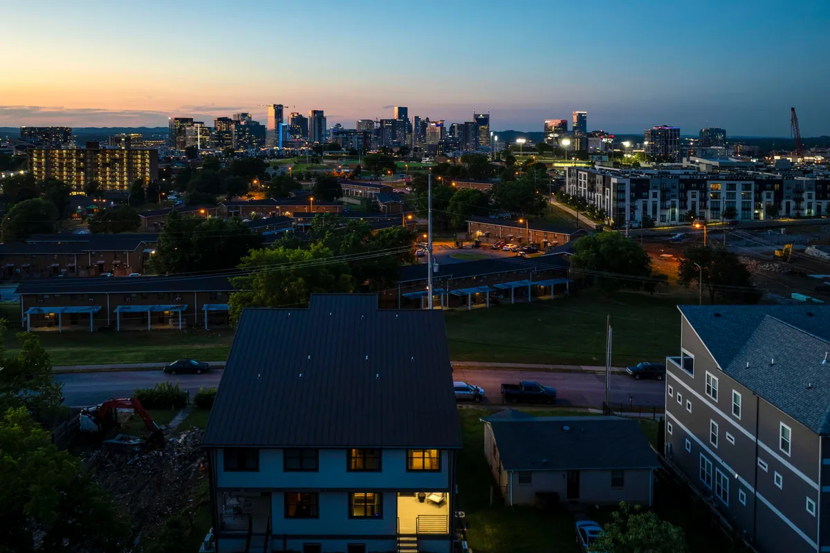 A sweeping city skyline view with illuminated windows, green spaces in the foreground, and a tranquil yet vibrant city atmosphere—suggesting the perspective one might enjoy from the rooftop deck during the evening.