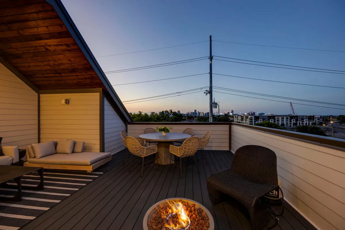 A cozy rooftop deck featuring a fire pit, outdoor dining table, and comfortable seating area. The vantage captures the city skyline beyond, with the setting sun casting a warm, golden hue over the space.